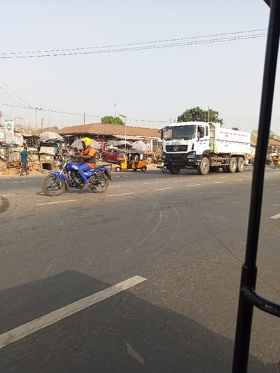 The image depicts a bustling street scene at a junction in Nigeria under a clear, bright sky. In the immediate foreground, a person wearing a yellow helmet, orange shirt, and blue trousers rides a blue motorcycle across the frame from left to right.

To the right, a large white Mercedes-Benz dump truck is prominently featured, displaying the company name "ABSOLUTE LOGISTICS LTD" on its side. Several yellow auto-rickshaws, locally known as "keke napep," are visible on the road and near the buildings, with a red car partially seen behind one of them.

The background reveals a lively market area characterized by various stalls with brown roofs and some covered by white or grey awnings. A person in a blue shirt is visible walking on the left side near some basic structures. Power lines are strung across the sky. The road is asphalt, marked with white lines and tire tracks. A dark metal frame, likely from the vehicle the photo was taken from, partially frames the right side of the image. The scene captures a typical sunny day with ongoing street and commercial activities.