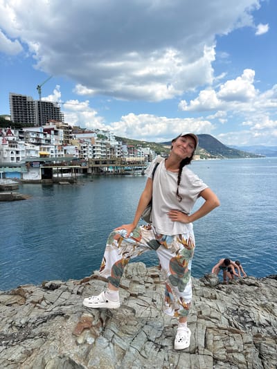 A woman stands on a rocky outcrop overlooking a body of water. She is wearing a light-colored t-shirt, patterned pants, a white cap, and white clogs. Her right hand is on her hip, and her left arm is bent with her hand on her leg. She is smiling and looking at the camera. In the background, there are buildings, mountains, and a cloudy sky. Two people are in the water to the right of the woman, appearing to climb out of the water onto the rocks. The image is taken in Kiparisnoe, Ukraine.
