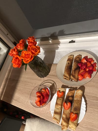 This indoor scene, identified as being in Arzberg, Germany, presents a top-down view of a light-colored wooden table surface adorned with breakfast or dessert items and floral decoration, positioned adjacent to a window.
In the lower foreground, a rectangular white plate holds three golden-brown rolled crepes. Each crepe is garnished with a single, heart-shaped slice of a red strawberry. To the left of this plate, a clear glass bowl contains multiple pieces of cut red strawberries.
In the upper midground, a circular, light-beige plate contains two additional golden-brown rolled crepes and a substantial pile of cut red strawberries. To the left of this plate, a dark-colored, fluted glass vase holds a bouquet of approximately six to seven vibrant orange roses with green stems and leaves.
The background consists of a white window frame, behind which a dark gray, fine mesh screen is visible. The screen features a distinct grid pattern. Strong, elongated shadows are cast across the wooden table surface from the window frame, the vase, and the plates, indicating a directional light source, possibly artificial or natural light from the window. No individuals are present in the image.