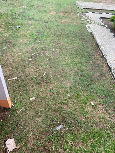 The image depicts a grassy area adjacent to a paved walkway in Sabo, Nigeria. The grass is patchy with areas of brown, dry vegetation interspersed with green. Scattered litter, including small pieces of plastic and paper, is visible on the grass. The paved walkway is constructed of light-colored interlocking blocks with a border of black and white painted concrete blocks. Part of a building with a grey and tan accent is visible in the lower right corner. The overall scene suggests a neglected or poorly maintained outdoor space. No people are visible.