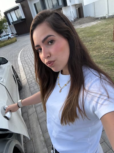 A young woman with long brown hair is holding the door handle of a white car. She is wearing a white t-shirt, a light-colored beaded necklace, and a white watch. The car is parked on a paved driveway or road in front of a building. The surrounding environment includes grass, trees, and other cars. Based on the architectural style of the buildings and overall setting, the location is likely Tlaxcalancingo, Mexico.