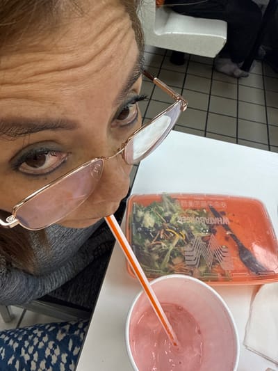 A person with eyeglasses appears to be sitting at a table with food and a drink. A partially eaten salad in a clear plastic container with a Whataburger logo sits beside a pink drink in a white cup with a straw. The person is wearing a gray top. The table is white and a section of blue patterned fabric is visible to the right. Tile flooring is in the background.
