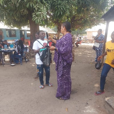 The image portrays an outdoor gathering in Assai Sabongida, Nigeria, under daylight conditions, with a prominent dirt ground surface. In the foreground, two adult women are the central focus. A woman on the left, wearing a white t-shirt, distressed blue jeans, and multicolored floral sandals, carries a black backpack with a blue-green object attached, and faces the woman on the right. The second woman wears a long purple patterned traditional dress and holds a dark handheld device, possibly a phone or recording instrument, directed towards the first woman.
To the left of the central figures, under a large tree, several individuals are seated at blue plastic tables, some with small white bowls on them. A blue-painted building is visible behind this group. To the right, other individuals are present, including a woman in a yellow t-shirt carrying a black bag and another in a blue and white patterned outfit holding a white bag. In the background, various simple structures, possibly market stalls, are visible, along with parts of a motorcycle. A fallen log or tree branch lies on the ground in the lower right portion of the scene.