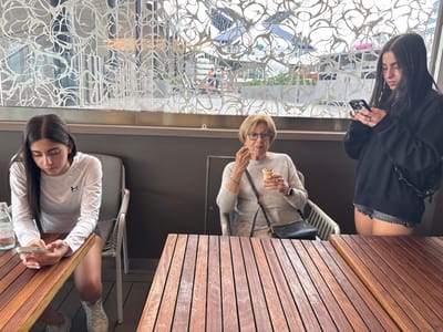 Three individuals are depicted on an outdoor patio or balcony in the City of Berry Islands, Bahamas. On the left, a young female with long dark hair, wearing a white long-sleeved Under Armour shirt and dark shorts, sits at a slatted wooden table, gazing at a smartphone held in both hands. A clear plastic water bottle is on the table beside her. Centrally, an elderly female with short light hair and eyeglasses, dressed in a light-colored long-sleeved top and a dark cross-body bag, is seated and consuming a chocolate-swirl ice cream cone with a spoon. To the right, a second young female with long dark hair, wearing a black long-sleeved hoodie and dark denim shorts, stands and focuses on a smartphone held in both hands. The background reveals a large window featuring an intricate white abstract screen, through which a blurred view of a waterfront area with distant boats and figures is visible.