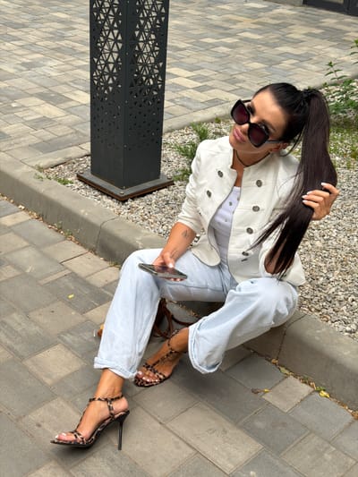 A woman is sitting on a sidewalk in front of a lamppost with a decorative geometric pattern. She is wearing a white jacket, a white top, light blue jeans, and leopard-print high-heeled sandals. She has dark hair pulled back into a ponytail, wears large sunglasses, and holds a smartphone in her hand. The ground is paved with interlocking bricks and gravel. The scene appears to be in an urban or suburban outdoor setting, possibly in Novaya Usman', Russia.