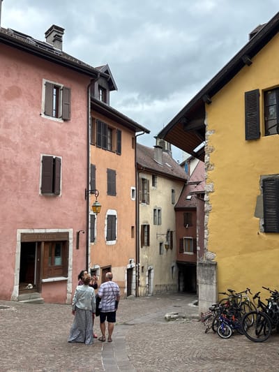 The image depicts a narrow, cobblestone street in Annecy, France, with colorful buildings on either side. A pink building is on the left, and a bright yellow building is on the right. Several people are visible: two individuals stand near the entrance of a shop on the pink building, one of whom is taking a photo of another person. A couple stands in the middle of the street, facing away from the camera, with the man wearing a plaid shirt and shorts, and the woman in a long skirt and jacket. A small white dog sits on the steps of the shop. Bicycles are parked against the yellow building on the right. The sky is overcast. The architecture suggests a historic European setting, consistent with Annecy's old town.