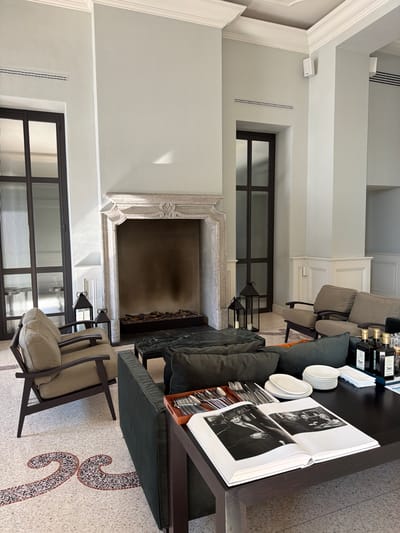 The image shows an indoor living room area in Pognana Lario, Italy, likely within a hotel or a high-end residential property. The room is elegantly furnished with a focus on comfort and style.
The focal point of the room is a large, ornate fireplace with a light-colored stone surround and a dark hearth. The fireplace is not currently in use, but the hearth contains decorative logs. Flanking the fireplace are tall, dark-framed windows with reeded glass, adding a touch of architectural interest.
In the foreground, a dark wooden coffee table is laden with items. An open coffee table book displaying black and white photographs is prominently featured. Beside the book are stacks of white plates, a cutlery set in a wooden tray, and several bottles of olive oil or similar liquid, possibly for decorative or functional use. A white napkin completes the arrangement on the table.
To the left of the fireplace, two dark, geometric lanterns with glass panels and lit candles are placed on the floor, adding a warm glow to the space.
The seating arrangements include two comfortable-looking armchairs upholstered in a neutral beige fabric, with dark wooden frames. These chairs are positioned to face the fireplace and coffee table. A dark green sofa with plush cushions is partially visible on the right side of the frame, and another dark green sofa is visible in the background behind the coffee table.
The floor is covered in a terrazzo-style mosaic with a decorative pattern featuring swirling shapes in shades of brown, beige, and pink, interspersed with small dark tiles. The walls are painted a light, neutral color, and architectural molding adorns the ceiling. The overall impression is one of sophisticated relaxation and luxury. There are no people visible in the image.