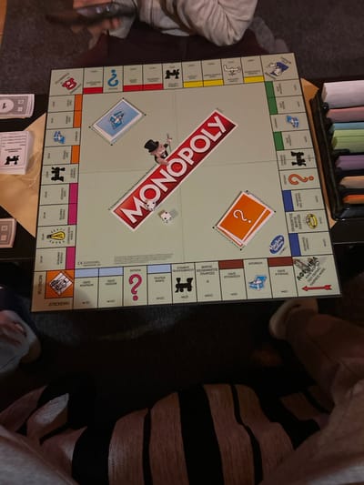 A Monopoly board game, featuring Greek text on its properties and cards, is laid out on a dark surface in the Gerakas Municipal Unit, Greece. Two white dice show values of 4 and 1, positioned near the red "MONOPOLY" logo. A black top hat game token is also visible near the logo. Stacks of blue Community Chest and orange Chance cards are in their respective designated areas on the board. A stack of white Monopoly money, with a visible '1' denomination, is on the far left. To the far right, a tray containing various colorful property deed cards is present. Three individuals are partially visible: one person at the top, wearing a grey long-sleeved garment, is holding and looking at a dark smartphone. Another person's lower torso and legs are visible at the bottom, wearing black and peach horizontally striped pants, with a light-colored shoe on their right foot. A third person's arm and knee are visible on the far left. The scene depicts an indoor setting, likely a casual gathering for playing the board game.