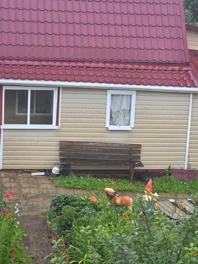 The image depicts a side view of a house in the city of Voskhod SNT (Frolovo), Russia. The house has a prominent red corrugated metal roof and beige siding. There are two windows visible on the house's facade, one larger with a white frame on the left and a smaller one with a white frame on the right, covered by white curtains. In front of the house, a weathered wooden bench sits on a brick pathway. Various garden decorations are scattered in the foreground, including a toy horse and a garden gnome with a red hat. Lush green vegetation and flowers are abundant around the pathway and in front of the house. The environment appears to be an outdoor setting, likely a backyard or garden area.