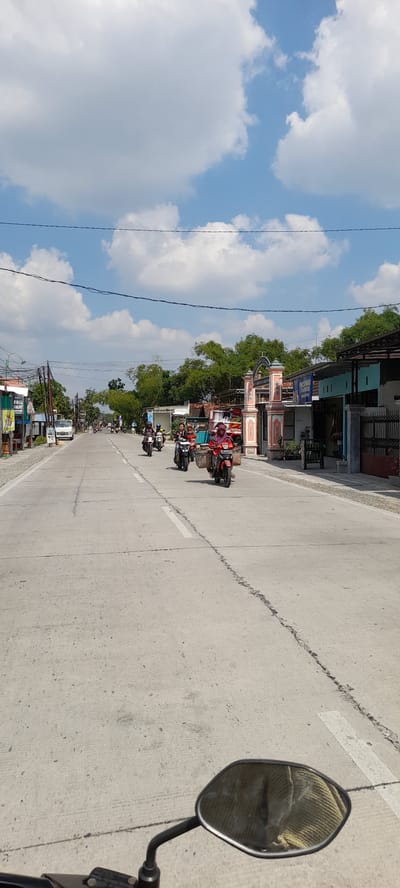 The image depicts a street scene in Grabagan, Indonesia, under a partly cloudy sky. The perspective is from a person riding a motorcycle, as indicated by the motorcycle's mirror in the foreground reflecting a textured object. The road is paved and shows signs of wear, with a dashed white line marking the center and a more solid white line on the right side.

Several motorcycles are traveling along the road in the same direction as the viewer. On the nearest motorcycle, a person wearing a pink headscarf and a pink top is carrying a large woven basket on the back. To their left, another person on a motorcycle is wearing a dark top and is also carrying something on the back. Further down the road, other motorcycles and riders are visible.

The right side of the road features a row of buildings with a distinctive archway that appears to be an entrance or a gate, decorated with orange and white patterns. Businesses are visible along this side, including one with a blue sign that reads "INDOMARET". Trees and foliage are present behind the buildings.

The left side of the road also has buildings and businesses, with utility poles and overhead wires spanning across the street. A white truck is parked on the side of the road. The environment appears to be a typical urban or semi-urban street with ongoing activity.