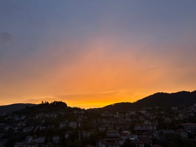 This image captures a panoramic view of Momchilovtsi, Bulgaria, at sunset. The sky is a gradient of deep blue at the top, transitioning to vibrant orange and yellow hues near the horizon, indicating the setting sun. The town is nestled on a hillside, with numerous houses with red-tiled roofs visible. The architecture suggests a traditional Balkan style. Lush green trees and dark, forested hills form the backdrop, contrasting with the illuminated sky. The overall impression is one of a serene, picturesque mountain village at dusk.