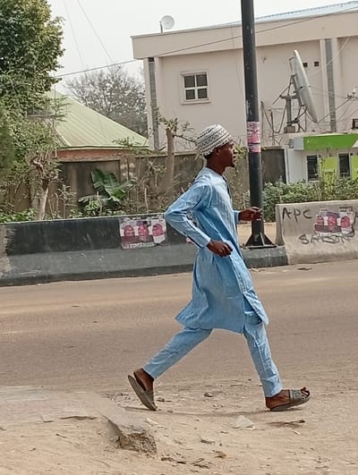 A dark-complexioned male walks from left to right in Darmanawa, Nigeria, on a dirt and broken concrete surface adjacent to a paved road. He wears a light blue long-sleeved tunic and trousers, a white and black patterned knitted cap, and brown open-toed sandals. His left leg is extended forward and his right arm is slightly forward, indicating mid-stride. A grey concrete barrier, affixed with several political posters including one marked "APC," separates the walking path from the road. To his right stands a black utility pole. In the background, a light-colored building with a flat roof and a small satellite dish is visible, alongside another building with a green sloped roof and a commercial-style building featuring green accents and a large ground-mounted satellite dish. Numerous trees and bushes are present throughout the background. The scene is illuminated by ambient daylight under what appears to be an overcast or hazy sky.