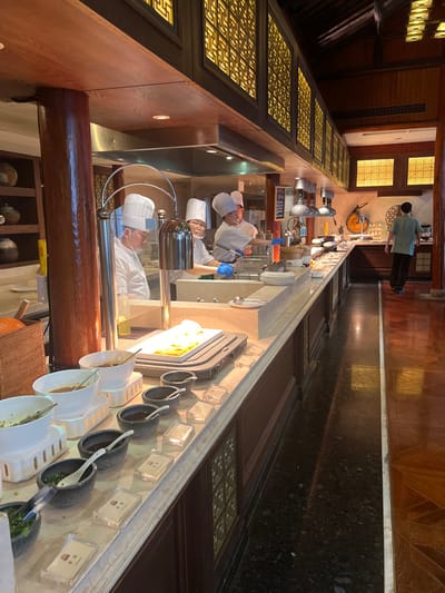 This image depicts an indoor dining area within a establishment in Tongxiang, China, featuring a live cooking and buffet station. The environment exhibits traditional Chinese architectural influences with dark wood columns, beams, and decorative overhead panels featuring backlit gold-toned geometric lattice patterns.

Four individuals identified as chefs are positioned behind a long, light-colored service counter. All chefs wear white chef jackets and tall white toque hats. Three are clearly visible wearing clear face shields, and two are equipped with blue protective gloves. The chef on the far right of this group is actively cooking at a hot station beneath a stainless steel exhaust hood, extending their right arm forward. Various pots, pans, and utensils are present on the stainless steel surface of the cooking area.

In the foreground, the buffet counter displays multiple food items and condiments. On the left, white ceramic bowls resting on white risers contain green and brown substances, likely sauces or salads. Several dark, small stone bowls containing dark sauces are arranged with white serving spoons. In front of these bowls, clear acrylic risers hold small white labels. Further along the counter, a large rectangular dish containing yellow food (potentially eggs) rests on a warming tray beneath two overhead stainless steel heat lamps with curved support arms.

To the right of the buffet, a dark, highly polished granite or stone floor strip separates the counter from a wider aisle featuring a polished, dark wooden floor with a parquet pattern. One individual, possibly a guest or staff member, is seen walking away from the camera along this wooden aisle. This person wears a light green or teal short-sleeved top and dark trousers, appearing to be in motion towards the far end of the room where a dark wooden console table with small white dishes and a decorative object is visible. The overall scene suggests a breakfast or lunch service in a well-appointed dining hall.