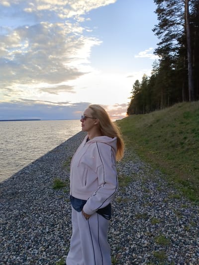 A woman stands near a body of water at sunset in Вичелово, Russia. She is wearing a light pink tracksuit with dark piping and sunglasses. Her blond hair is long and flowing. To her left is a body of water with land visible in the distance. To her right is a grassy embankment leading to a line of dark evergreen trees. The ground she is standing on is covered in small, light-colored rocks. The sky above is cloudy with the sun setting.