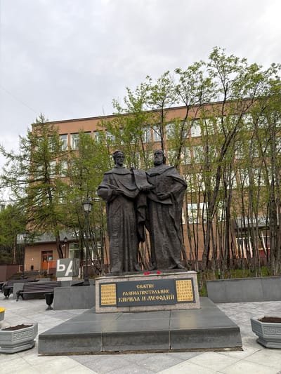 The image shows a monument to Saints Cyril and Methodius in Murmansk, Russia. The bronze statue features the two saints standing together, clad in robes. In front of the statue, there is a stone plaque with text in Cyrillic. Behind the monument, there are trees with young green leaves, partially obscuring a light brown building with multiple windows. The sky is overcast. The monument is set on a paved area, and a park bench is partially visible to the right.