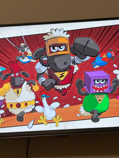 This is a close-up, tilted, eye-level shot of a digital illustration displayed on a screen. The illustration depicts a chaotic scene with several cartoonish characters and objects against a vibrant, textured background. The background features diagonal red and orange stripes, suggesting a stage or a dynamic environment.

The main subjects are anthropomorphic characters, primarily sheep and some other creatures rendered in a bold, graphic style. On the left, a character with a dark grey, helmet-like head and a yellow and red chest piece resembling a robot is partially visible. It has large, circular eyes with pupils. To its right, another character, a purple box with multiple eyes and a red, dotted mouth, is shown with black limbs. Next to it, a green character with a yellow triangular hazard symbol is also depicted with black limbs.

In the upper part of the image, a red character with blue and yellow accents, resembling a stylized cow or bull with horns, is suspended in the air. A white cartoon chicken with yellow feet is falling or in motion, with a small blue and red rooster-like character near its head. Below these, several sheep characters are prominently featured. One sheep, wearing a golden crown with red circles and white fluff, is on the right side of the frame, its body is mostly obscured by the screen's edge. Another sheep, with a red body and black ears and eyes, is in the lower-middle section, seemingly interacting with some laser-like effects.

Feathers are scattered throughout the scene, adding to the sense of action and disarray. The overall tone of the illustration is energetic and playful, with bright colors and exaggerated features. There is no visible text in the image. The lighting suggests it's a screen display, possibly in an indoor setting, with the ambient light creating reflections on the screen's surface. No specific time of day or weather conditions are indicated. The location context of Berezhany, Ukraine, is not visually represented in the illustration itself.