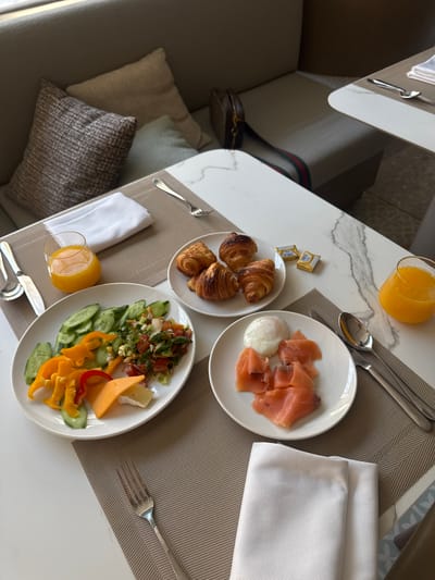 The image depicts a breakfast setting on a white table, possibly in a hotel or apartment in El Gouna, Egypt. Two white plates are visible; one contains smoked salmon and a poached egg, while the other features a salad composed of cucumbers, bell peppers, cheese, and a chopped vegetable mix. Also present are croissants on a smaller white plate, packets of butter, and two glasses filled with orange juice. Silverware (forks, spoons, and knives) is neatly arranged. A white linen napkin is positioned near each plate. A section of a sofa with decorative pillows is visible in the background. A handbag is also partially visible. The scene suggests a relaxed and comfortable breakfast setting.