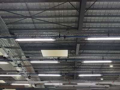 The image shows the interior of a large industrial space, likely a warehouse or factory, with a high ceiling constructed of corrugated metal panels supported by a grid of steel beams. Rows of fluorescent lights hang from the ceiling, illuminating the area. Metal cable trays are suspended alongside the lights, carrying wiring and infrastructure. A rectangular skylight is visible, providing natural light. The overall impression is of a functional, utilitarian space.