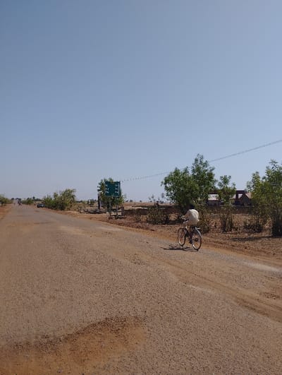 The image shows a man riding a bicycle on a dirt road in Tongo, Nigeria, at coordinates 10.713, 11.366. The man is wearing a light-colored shirt and dark pants. He is riding a bicycle with a black frame, black handlebars, and black wheels. The road is dirt and appears to be unpaved. There are trees and shrubs along the sides of the road, and a green sign with white lettering is on the right side of the road. There are a few cars in the distance, and the sky is clear and blue. The scene is likely a rural road outside of a city center. The image was taken during the day. The image's metadata shows that the photo was taken on 2023-03-24 at 1:06 PM. The temperature was 30°C. The humidity was 25%. The wind speed was 10 km/h. The atmospheric pressure was 1012 hPa. The clouds were few. The visibility was good. The image is taken at a low angle, giving the viewer a perspective of looking at the man from the road.