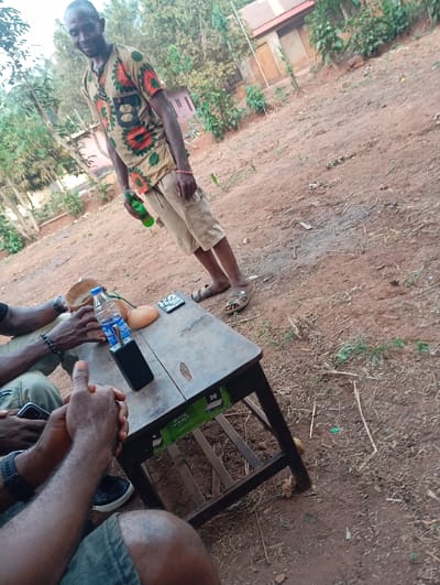 An outdoor scene features a man in a patterned shirt and cargo shorts holding a beer bottle. He stands near a small, weathered table where at least two other people are seated. The table holds a water bottle, what appears to be a gourd, and a black electronic device. The ground is dirt and foliage, with a building visible in the background.