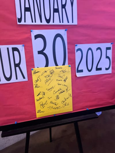 The image displays an indoor setup featuring a red surface, likely a large board or table, adorned with white cards pinned by blue thumbtacks. These cards spell out a significant date: "JANUARY 30 2025". A partially visible card to the bottom left shows "UR", suggesting it's part of a day of the week, such as "THURSDAY".

A yellow card, also pinned with blue thumbtacks, is placed next to the "30" card. This yellow card is covered with numerous handwritten signatures and names in black ink. Visible names include "Heathere", "Denny", "Paul Konter", "Conchita", "Billie", "Kellen", "Andy", "Melanie", "Brod", "Sox", "Chao", "Evelyn", "Leven", and "Allen".

The scene suggests an event or announcement related to January 30, 2025, possibly a future event, an important deadline, or a milestone being acknowledged by a group. The collective signatures imply participation, agreement, or a shared commitment towards this date. The setting is likely an office, community center, or event space in South Brisbane, Australia. The image does not provide clues about the time of day or specific emotions, but the collaborative nature of the signatures points to a collective purpose.