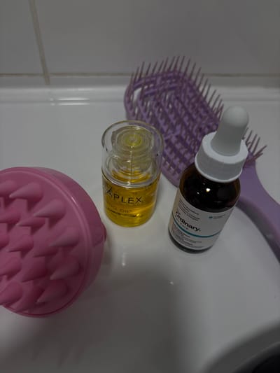 The image shows three items on a white countertop. The items are a pink hairbrush, a small bottle of yellow oil labeled "PLEX" and a small bottle of brown liquid labeled "The Ordinary." The location of the photo is Ramla, Israel, based on the coordinates provided. The bottles appear to be in focus and the hairbrush is slightly blurred. The image is well-lit and the background is a white tile wall.