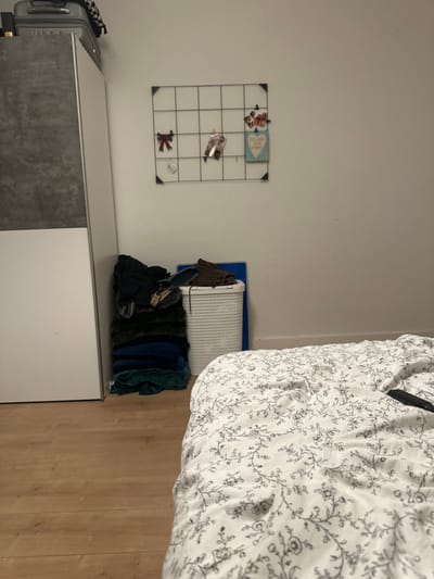 This image depicts an interior room, specifically identified as a bedroom located in Montreux, Switzerland. No individuals are present, and no active interactions or actions are occurring.
The foreground on the right displays a bed covered with a white duvet featuring a repeating pattern of dark, stylized floral or vine motifs. A black object, consistent with a remote control, rests on the duvet. The flooring throughout the visible area is light brown laminate wood.
On the left side of the room, a tall, two-toned wardrobe or cabinet stands against the wall, presenting a white lower section and a grey upper section. A dark grey hard-shell suitcase, topped with a black and white checkered fabric item, is positioned on top of the wardrobe. Adjacent to the wardrobe, a stack of dark-colored textiles, primarily in shades of green and blue, is neatly piled on the floor. Beside this stack, a white plastic laundry basket with a textured surface holds additional items, and a flat, rectangular blue object (possibly a mat or board) leans against the wall behind it.
Centered on the light-colored wall above the laundry area is a black metal grid-style wall organizer. Attached to this grid are several decorative items: a red plaid bow, a small white hanging decoration, a clipped photograph or card, and a light blue heart-shaped sign bearing the text "FOLLOW YOUR HEART."