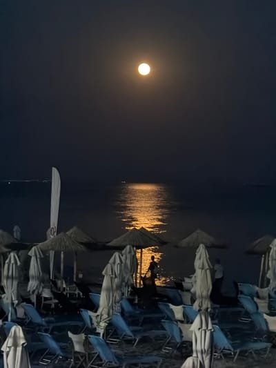 The image depicts a nighttime scene at a beach in Nea Kallikratia, Greece, illuminated by a full moon. The moon is prominently positioned in the dark sky, casting a golden reflection on the calm sea. The foreground shows a row of closed beach umbrellas and lounge chairs, suggesting the beach is unoccupied or has been prepared for the night. A white flag is visible on the left side of the image, and two faint figures are seen standing by the water's edge, silhouetted against the moonlight. The overall atmosphere is serene and tranquil.