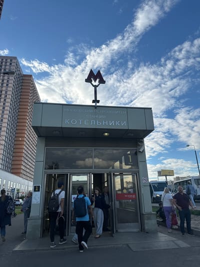 A low-angle shot captures the entrance to the Kotelniki metro station in Moscow, Russia, under a bright blue sky filled with wispy white clouds. The metro station building is modern with glass doors and metal accents, featuring the distinctive Moscow Metro logo.
Several people are visible, some queuing to enter the station and others walking by. A man in a dark red shirt and jeans stands near the entrance, while a woman in a white top and purple pants is next to him. Other individuals, some with backpacks, are seen entering or near the glass doors.
Text on the building reads "МОСКОВСКИЙ МЕТРОПОЛИТЕН" and "СТАНЦИЯ КОТЕЛЬНИКИ", identifying it as the Moscow Metro Kotelniki station. A sign near the entrance displays "1 Вход" (1 Entrance). A red sign on the glass door indicates "Нет входа" (No Entry) with a symbol of a person with a line through them.
In the background, a tall, modern building with a patterned facade and a skyscraper stand against the sky, suggesting an urban environment. The overall scene appears to be during daylight, possibly late morning or afternoon, given the bright sunlight casting shadows. The weather is fair with a partly cloudy sky.