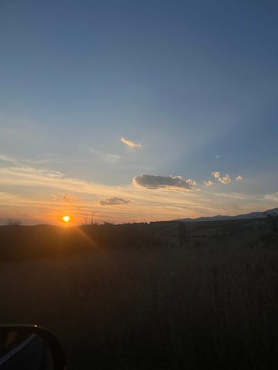 The image depicts a rural landscape at sunset, with the sun visible as a bright orb near the horizon. The sky is a gradient of blues and oranges, with scattered clouds reflecting the sunlight. A silhouette of hills or mountains can be seen in the distance. The foreground is dominated by tall grass and vegetation, casting the immediate area in shadow. A portion of a vehicle, likely a car, is visible in the lower left corner. The scene appears tranquil and natural, with no specific structures or activity indicating an urban environment. The reference to Domlyan, Bulgaria, suggests the photograph was taken in or around that location.