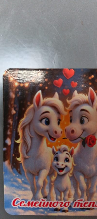 The image shows a card with a cartoon illustration of three white horses, two adults and one foal, in a snowy, festive setting with glowing lights and red hearts. The adult horses appear to be a couple, with one holding a red rose in its mouth. The foal stands between them, smiling. At the bottom of the card, text in Russian reads "Семейного тепла" which translates to "Family warmth". This card is likely a decorative item or a greeting card. There is no indication of the city of Tikhvin, Russia in the image itself.