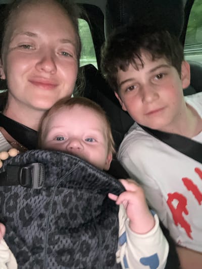The image depicts a woman, a young boy, and an infant in a car, likely captured as a selfie. The woman is seated in the front passenger seat, while the boy sits behind her. The infant is positioned in a black and grey leopard print baby carrier worn by the woman. The woman is smiling and looking at the camera, the boy appears to be looking away from the camera and the infant is looking directly at the camera. The boy is wearing a white t-shirt with a large red graphic on the front, and the infant is wearing a white long-sleeved shirt with a blue pattern. The woman is wearing a cream-colored sweater and a gold necklace with a wooden bead pendant. The woman's hair is light blonde and pulled back. The boy's hair is dark brown and brushed forward. The infant's hair is light red and looks fluffy. The environment is a car interior, with dark-colored seats. The image appears to be taken at night, as there is limited light inside the car. The coordinates provided, 45.653 latitude, 8.74 longitude, place the image location in the town of Cardano al Campo in the province of Varese in Lombardy, Italy.