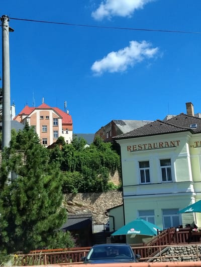 The image shows a street scene in Vimperk, Czechia, on a sunny day. In the foreground, a dark-colored car is partially visible, parked behind a wooden fence. A bright green umbrella with the Pilsner Urquell logo is open next to a restaurant building. The building, painted a light yellow, has "RESTAURANT" written in large brown letters above its windows. Behind the restaurant, a stone wall supports a hillside covered in lush green trees and vegetation. Further back, a multi-story building with a red roof is visible, and to its right, another building with a dark tiled roof. The sky is clear blue with a few white clouds. A tall concrete utility pole with wires stretches across the upper left portion of the frame.