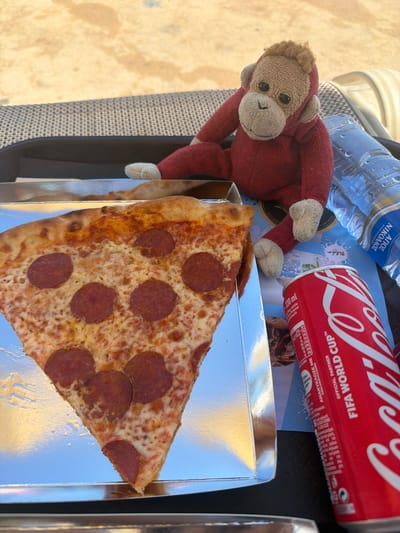 This media file shows a close-up overhead view of a pepperoni pizza slice in a silver foil tray, accompanied by a red Coca-Cola can and a bottle of water. A small, plush monkey toy in a red outfit is placed next to the pizza and can. The Coca-Cola can is decorated with "FIFA WORLD CUP™" text. The water bottle has Greek text, which translates to "Saint Nicholas Natural Mineral Water". The scene appears to be outdoors, possibly on a patio or table, with a light-colored, textured surface visible in the background. The lighting suggests it is daytime, and the overall mood is casual, implying a snack or meal break. The setting does not provide strong clues to pinpoint a specific location within Trachoni, Cyprus, beyond the general outdoor context.