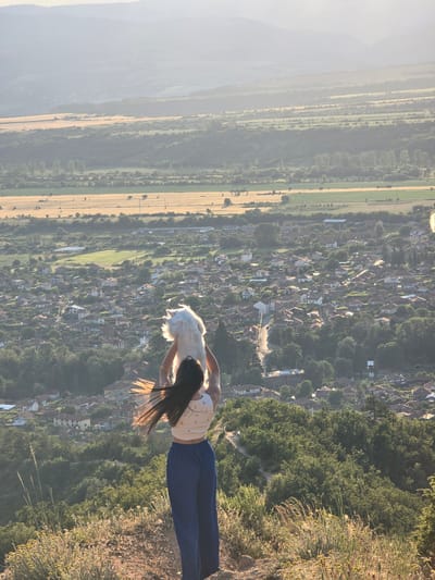 The image presents a high-angle view overlooking the village of Stob, Bulgaria, and the surrounding valley, likely captured during late afternoon or early evening. In the foreground, a person stands on a dry, grassy hilltop, facing away from the camera. They have long, dark hair, which is styled backward and appears to be in motion, possibly by wind. The individual wears a light-colored, subtly patterned crop top and dark blue, wide-legged trousers. Their arms are raised overhead, holding a small, white, fluffy dog that is also oriented to face the distant landscape.
Below the hilltop, the village of Stob is visible, characterized by a dense cluster of buildings, primarily with reddish-brown tiled roofs, interspersed with numerous green trees. A road winds through the village. Beyond the village, the valley floor exhibits a mosaic of agricultural fields, some appearing golden and others green, along with patches of forest and scattered structures. Distant, hazy mountains form the horizon under a bright, diffused sky.