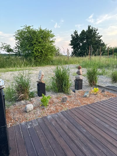 The image depicts an outdoor scene with a wooden deck in the foreground, running diagonally from the bottom right corner towards the top. Beyond the deck, a gravel and small stone area is visible, interspersed with patches of tall green grass and some small plants. Two black rectangular objects, possibly planters or decorative elements, are placed among the grass, each topped with a stack of smooth, grey stones. One of these stacks features a round, reddish-brown stone on top. In the background, a paved path or road is visible, bordered by more grass and foliage. Further back, large trees with dense green leaves are present. The sky is a clear blue with scattered white clouds, suggesting it is daytime, possibly late afternoon or early evening due to the soft lighting. The overall setting appears to be a garden or outdoor living space, possibly in a rural or semi-rural location, given the natural elements and open sky. There is no visible text in the image.