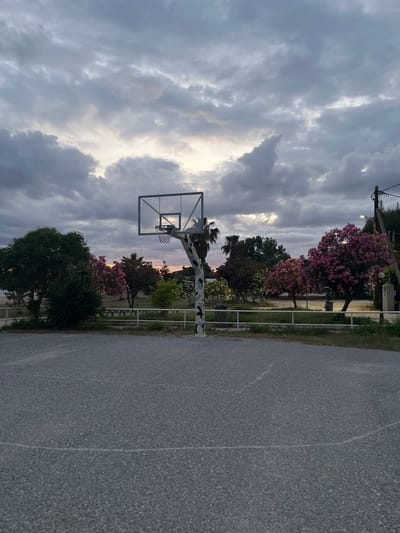The image depicts a basketball court with a hoop and backboard in the foreground. Behind the court, there is a line of trees and flowering bushes, with a fence separating the court from the greenery. In the background, the sky is filled with dramatic, cloudy formations, suggesting either dawn or dusk. The overall scene appears to be outdoors, possibly in a park or recreational area. The location is identified as Gerakini, Greece. There are no people visible in the image.