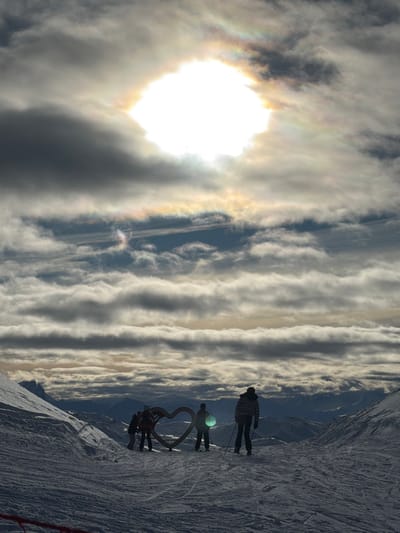 The image displays a vibrant snowy mountain landscape under a dynamic sky, oriented horizontally despite the image being provided in a vertical (sideways) presentation.
On the left side, a brilliant, almost blown-out sun shines through hazy clouds, creating a strong solar halo or iridescence. This phenomenon manifests as rainbow-like colors, particularly vivid directly around the sun and extending into the surrounding clouds. Darker cloud formations partially obscure the sun, casting shadows and creating varied light effects.
Stretching across the mid-ground and toward the right, the sky is filled with numerous long, horizontal cloud bands, creating a striking striped pattern. These clouds vary in density and brightness, with some appearing bright and illuminated, while others are darker, and patches of blue sky are visible in between.
The right side of the image features a snow-covered mountain slope, appearing steep in the foreground. Distinct ski tracks are visible on the snow, indicating recent activity. In the distance, faint outlines of other mountain ranges or hills can be seen, softly blending into the sky.
Several human figures are visible on the snowy slope, silhouetted against the bright sky and clouds. At the upper right, a solitary figure stands upright, holding ski poles and wearing what appears to be a plaid jacket. Below and slightly to their left, another person stands with ski poles, notable for a distinct bright, possibly glowing, green or blue spherical object attached to their person or equipment. Further down the slope and to the left of these two figures, a prominent dark, heart-shaped structure or sculpture stands on the snow. Around this heart sculpture, several more dark figures are gathered, suggesting a group stopping to admire the view or take photos.
The overall scene suggests a recreational setting, likely a ski resort, where people are enjoying the mountain environment. The low-angle sunlight, silhouetted figures, and the quality of light indicate either early morning or late afternoon. The presence of the solar halo points to specific atmospheric conditions, possibly involving ice crystals in the clouds. There is no visible text in the image.