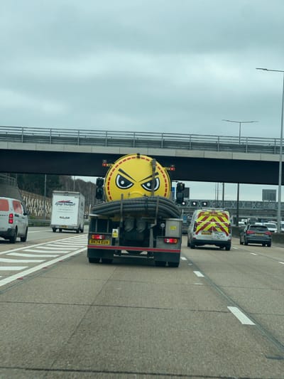 A forensic, matter-of-fact description of the scene follows:
The image depicts a multi-lane asphalt motorway under an overcast sky, observed from the perspective of a vehicle in motion. The prominent subject in the foreground is the rear of a yellow tanker truck, registered in the UK with license plate RK74 EUV. The rear face of the cylindrical tank is adorned with a large, stylized cartoon angry face decal in black and white. Below the tank, multiple thick black hoses are coiled horizontally across the rear flatbed of the truck. The truck's rear features red tail lights, amber indicators, and various warning decals.
Multiple vehicles are present on the motorway. To the left of the tanker, a white van is partially visible in an adjacent lane. Further left and behind, a white box truck with "Lounge Transport" text and contact details is observed. To the right of the tanker, a white utility van with "HIGHWAY MAINTENANCE" marked on its rear, along with yellow and red chevron reflective tape, is in another lane. This utility van also displays "nets" branding and has a partial registration plate (MJ6...LJK). Behind the utility van, a dark grey Audi SUV is visible.
The road surface is asphalt, delineated by white solid and dashed lane markings, and white hatched markings are present on the left side, possibly indicating a gore area or a narrowed lane. An overhead concrete bridge structure with metal railings spans the motorway. Streetlights with distinctive T-bar crossarms are visible along both sides of the road. In the far left background, a wooded area borders a patterned retaining wall or sound barrier.
The location is identified as the A3 northbound, within or immediately adjacent to the city of Guildford, Surrey, United Kingdom, characterized by the specific bridge and multi-lane highway infrastructure evident in the image.