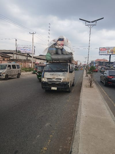 The image depicts a busy, overcast daytime scene on what appears to be an urban or semi-urban street. The perspective is from the side of the road, looking forward along the direction of traffic.
In the foreground and center, a white or light-colored utility van or small truck is heavily overloaded with goods on its roof rack. The vehicle itself appears weathered, with visible dirt or discolored patches on its front and sides. Its front grille and headlights are visible, and the windshield reflects the sky. The load on top consists of numerous large sacks or bags, predominantly white and grey/blue, piled high and seemingly secured with ropes or tarps. The load significantly exceeds the height of the vehicle's cabin.
The vehicle is traveling on a two-lane asphalt road. Yellow dashed lines are faintly visible on the road surface, indicating lane divisions. To the right of the road, a raised concrete curb separates the main thoroughfare from a narrow concrete sidewalk or median, which shows some minor debris or sparse vegetation.
Along the left side of the road, a variety of buildings, possibly shops or residential structures, line the street in the mid-ground and background. Multiple utility poles with a dense network of power lines stretch across the sky. Two prominent billboards are visible:
1. In the mid-ground, a large billboard displays an advertisement featuring the brand name "LAZIZ" in large red letters, with "QUALITY" and "TASTE" written below it. The billboard also shows images of food products, appearing to be fried chicken pieces, french fries, and soft drinks.
2. Closer to the foreground, another billboard stands with what appears to be a graphic and some text, which is largely unreadable but includes script that resembles Arabic or a similar language.
Other vehicles are visible on the road: a dark-colored car is partially seen directly behind the overloaded truck, and another light-colored van or small bus is parked or moving further down the road on the left side. Traffic appears moderate.
The sky is uniformly overcast with grey clouds, indicating a cloudy day, but the scene is well-lit, characteristic of daytime. Modern streetlights with twin rectangular fixtures are mounted on tall poles along the roadside.
Visible text includes:
* On the billboard: "LAZIZ", "QUALITY", "TASTE".
* On the other billboard: Some unreadable text, possibly in Arabic script, and "FRESH".
* On the overloaded truck's side: Partially visible text, possibly "EXPRESS" or similar.
* On the license plate of the dark car behind the truck: "LAGOS" is partially visible.