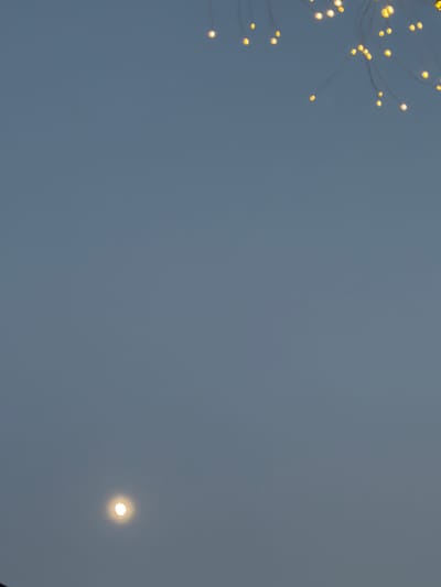 The image shows a sky with a faint, diffused light, possibly indicating dusk or dawn. In the upper left portion of the frame, a cluster of small, bright yellow lights is visible, appearing to be part of a decorative light installation. These lights are strung on thin, delicate wires, creating a wispy, ethereal effect. In the lower right quadrant of the image, a single, bright, circular light source is present, which could be the moon or a distant celestial body. The sky itself is a muted, grayish-blue, suggesting overcast conditions or the dimming light of twilight. The overall mood of the image is serene and somewhat mysterious, with the contrasting lights against the soft sky creating a sense of quiet beauty. The location context is Kamianka-Buzka, Ukraine, though no specific landmarks are visible to confirm this. No people are visible in the image, and there is no discernible text.