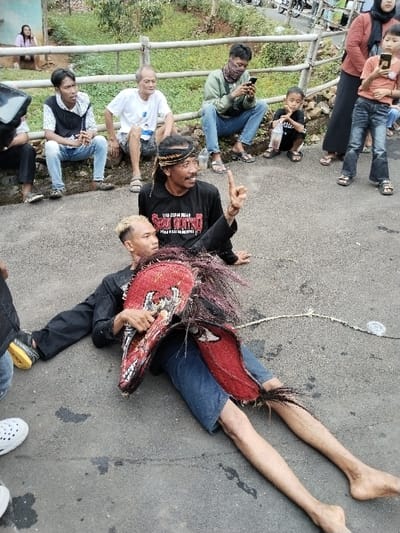 A man with a headband sits behind a person wearing a large, red, feathered mask and lying on the ground. The masked person wears a black shirt and blue shorts, with very thin legs. A rope is attached to the mask. Several people are seated or standing in the background, including a man looking at a phone, children, and a woman in a hijab. The setting appears to be a rural road or paved area with a fence and greenery in the distance.
