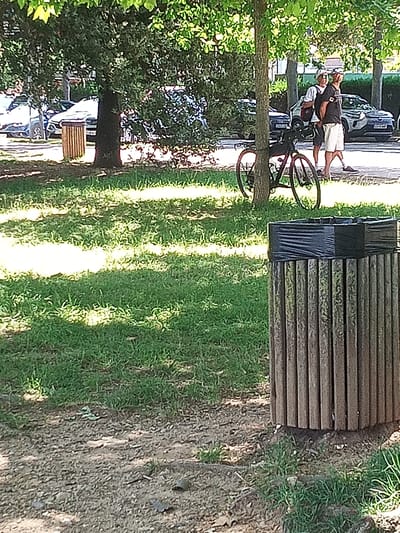 The image depicts a park setting with a paved pathway, grass, and trees. In the foreground, a wooden trash receptacle with a black liner is visible. To the left, a bicycle is leaning against a tree. Further back, two men are standing near parked cars. The overall scene suggests a public outdoor space. It is not possible to determine the location in Banyoles, Spain based on the visual information provided.