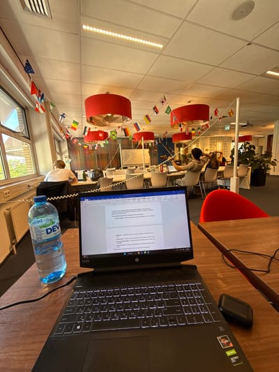 This is an indoor shot of a modern office or study space, possibly a university library given the presence of multiple people working. The scene is captured from a high angle, looking down and across a room adorned with a string of small national flags, creating a festive and international atmosphere. The lighting suggests it's daytime.
In the foreground, a laptop is open on a wooden desk, displaying a document that appears to be academic or professional text, as indicated by the "AaBbC" formatting tools visible on the screen. A water bottle with a blue cap, labeled "DEV Om" and "НАТУРАЛНА," is positioned near the laptop. The laptop itself has illuminated keys, suggesting it's actively in use.
Beyond the immediate workspace, several individuals are seated at tables, seemingly engaged in their studies or work. They are mostly facing away from the camera, their backs to the viewer as they focus on their own devices or materials. In the background, there are more tables, chairs, and some decorative elements like red cylindrical lamps and potted plants. The overall ambiance is one of quiet concentration and productivity, typical of a shared workspace. The string of flags hints at a diverse community or an event celebrating international diversity, common in academic institutions in locations like Enschede, Netherlands. The visible text on the laptop screen includes phrases like "Transactional leadership," "Transformational leadership," and "motivation," suggesting the topic of the document relates to leadership studies.