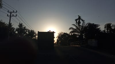 The image depicts a scene at dusk or dawn on a rural road in Kruengseupeng, Indonesia. The sun is low in the sky, creating a silhouette effect of the surrounding elements. A large truck, also silhouetted, is visible on the left side of the road, appearing to be stationary or moving slowly. The road itself is dark and appears to be asphalt. To the right of the truck, there is a bridge or elevated structure with a railing. Beyond the bridge, more silhouetted trees, including palm trees, are present. A person is faintly visible on a motorcycle on the bridge. In the background, a utility pole with multiple wires stretches across the sky. The sky is a gradient of pale blue and yellow, indicating the presence of the sun. There are no other discernible people or vehicles in the immediate vicinity. The overall atmosphere is serene and rural.