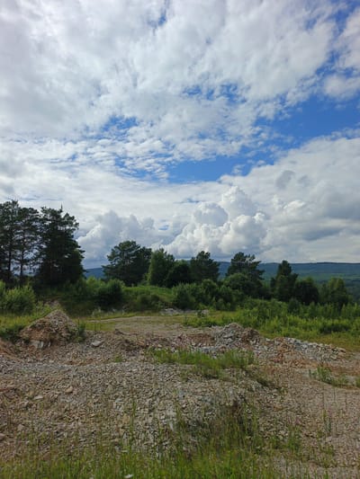 This media file captures a scenic view of a landscape with a focus on a rough, rocky terrain in the foreground and a lush forest and hills in the background under a cloudy sky. The foreground is dominated by piles of stones and gravel, suggesting a possible construction site or a natural area that has been disturbed. The texture of the rocks is varied, with some appearing sharp and angular, while others are more rounded.
Beyond the rocky foreground, the scene transitions into a dense, green forest with various types of trees, including tall pine trees on the left. The vegetation appears healthy and vibrant, indicative of a natural, possibly rural setting. In the distance, rolling hills covered in trees stretch towards the horizon.
The sky is a prominent feature, filled with dramatic, fluffy white clouds that contrast with patches of bright blue sky. The lighting suggests it is daytime, likely late morning or early afternoon, given the intensity of the light and the shadows cast. The weather appears to be mixed, with the clouds hinting at potential changes, but currently offering a visually striking backdrop.
There are no people or identifiable man-made objects in the scene, apart from the gravel piles, which could be interpreted as remnants of human activity. The overall mood is one of natural beauty and ruggedness, with a sense of tranquility despite the unfinished nature of the foreground.
The location context provided, Urtiushka, Russia, places this scene in a specific geographical area, suggesting a natural landscape typical of that region. There is no visible text within the image.