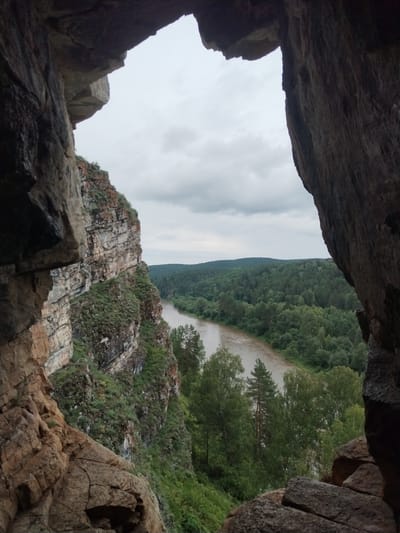 The image is a photograph taken from within a cave opening, looking out at a natural landscape. In the foreground, dark, textured rock formations frame the view. To the left, a steep, layered cliff face is covered in patches of green vegetation. A winding river, with murky, brown water, flows through a valley. The valley is densely forested with various shades of green trees, and rolling hills can be seen in the distance under an overcast sky. The location is identified as Idrisovo, Russia. There are no people or human activity visible in the image; it is a scene of natural scenery.