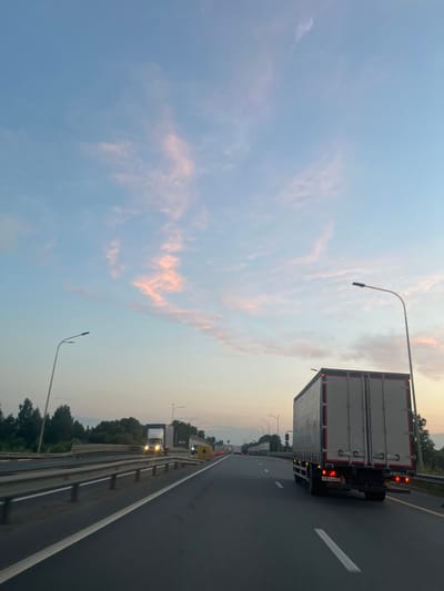 The image depicts a highway scene at dusk, with a clear sky showing streaks of pink and orange clouds against a pale blue backdrop. A large, gray semi-trailer truck with red taillights is in the foreground, traveling on the right lane of a multi-lane highway. The truck's license plate is partially visible, reading "K 901BE 37". Several other vehicles, including trucks and cars, are visible in the distance on the highway. The roadside features trees and vegetation, as well as streetlights and guardrails. The overall environment suggests a journey on a major road outside of a densely populated urban area, possibly near the city of Konino, Russia, given the context. The lighting and atmospheric conditions indicate it is either sunrise or sunset.