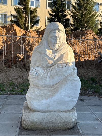 The image displays a large, light-colored stone statue situated outdoors in Aksay, Kazakhstan. The primary subject is a statue, likely carved from concrete or a similar light-hued stone, depicting a person, possibly a woman, with a serene or somber expression. The figure wears a head covering, resembling a veil or shawl, and is enveloped in flowing drapery, characteristic of religious or commemorative artwork. The statue appears to be in a seated or kneeling position, with hands subtly visible beneath the folds of the garment.

The statue rests on a roughly textured pedestal and is positioned next to a paved walkway constructed from grey concrete slabs. Behind the statue, a dark metal fence with decorative scroll patterns acts as a barrier, separating the paved area from a mound of brown earth or dirt. Further in the background, dark green evergreen trees are visible, partially obscuring a light-yellow or beige multi-story building featuring numerous rectangular windows.

The scene is illuminated by bright natural sunlight, indicating it is daytime, likely morning or late afternoon given the prominent, sharp shadows cast by the statue and the fence. The clear lighting emphasizes the textures of the stone carving and the surrounding environment. No visible text is present in the photograph. The setting suggests a public space, possibly a park or an institutional ground, conveying a sense of quiet solemnity.