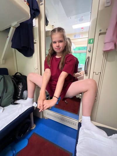 A young girl with long blonde hair, styled in braids, is positioned in a seated, kneeling pose within a train compartment. She is wearing a dark red t-shirt and white socks. Her expression is direct and engaging, looking towards the camera. Her arms are crossed and resting on her knees. The compartment features typical train interior elements such as overhead luggage racks, a window showing greenery outside, and bunk beds. One bunk is covered with white bedding, suggesting it is occupied or prepared for sleep. Dark blue fabric is visible on a lower bunk or seat, and several bags, including a green backpack, are stowed in the compartment. The lighting suggests it is daytime. The overall atmosphere is casual and suggests a journey in progress.